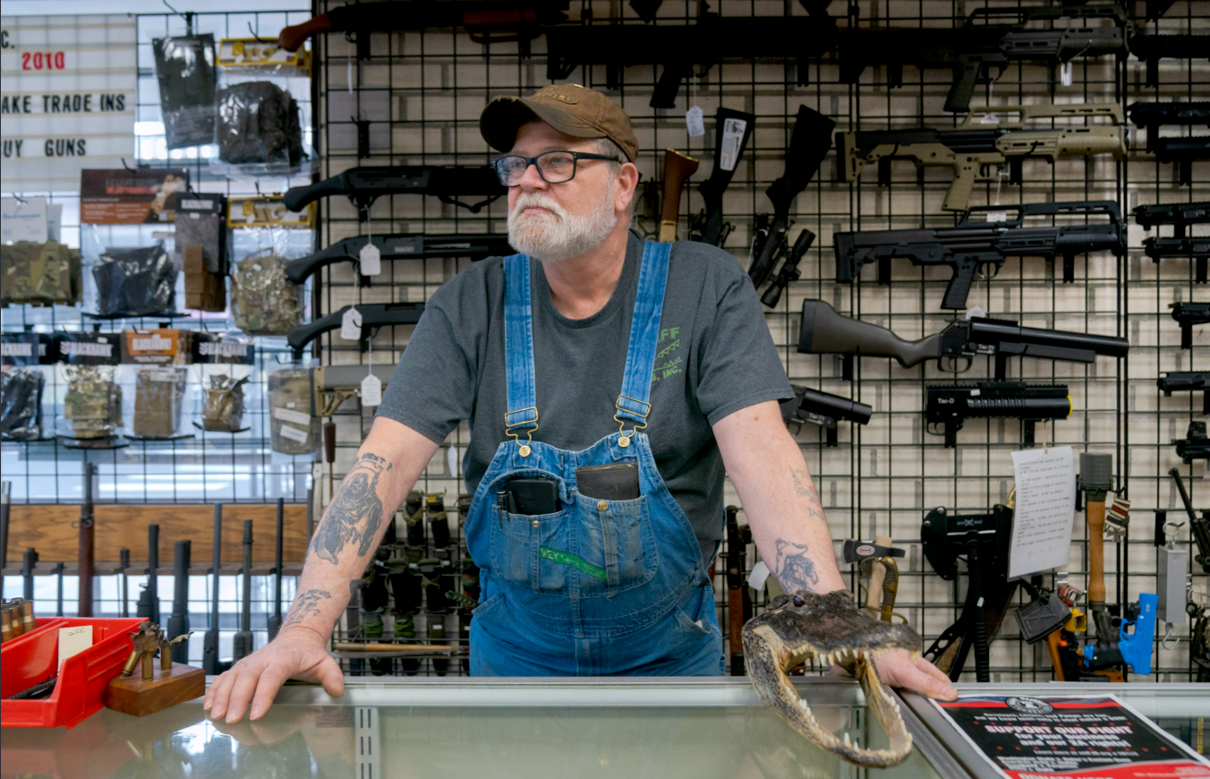 Gun store employee standing behind a display counter with firearms on the wall