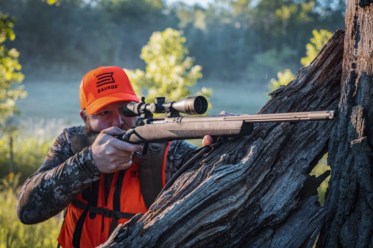 Hunter wearing blaze orange Savage Arms gear aiming a scoped bolt-action hunting rifle supported against a fallen tree in a wooded setting.