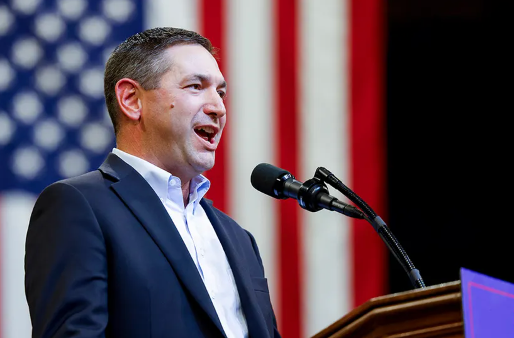 Montana Attorney General Austin Knudsen speaking into a microphone at a podium with an American flag in the background.