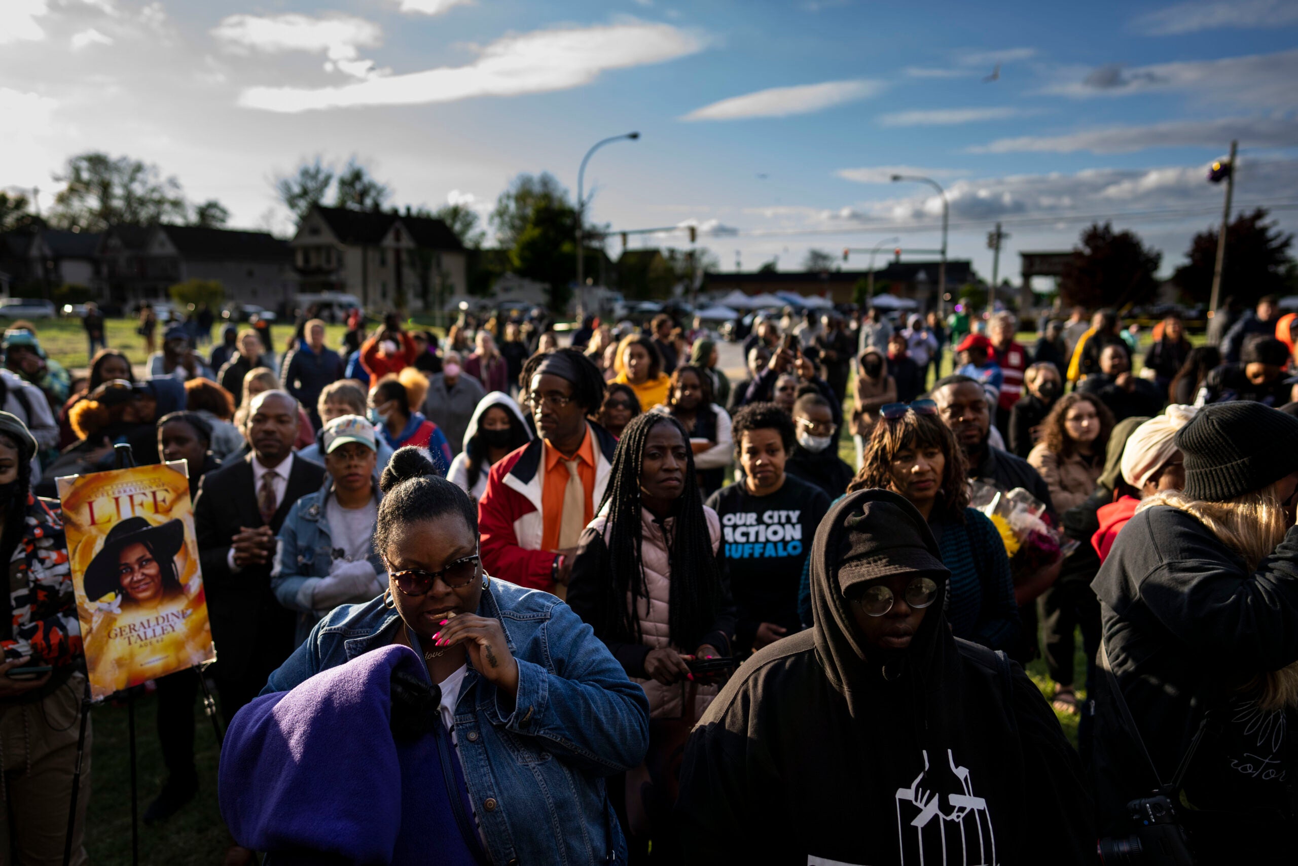 A large, diverse crowd gathers outdoors in Buffalo, New York, with several attendees holding flowers and memorial photographs.