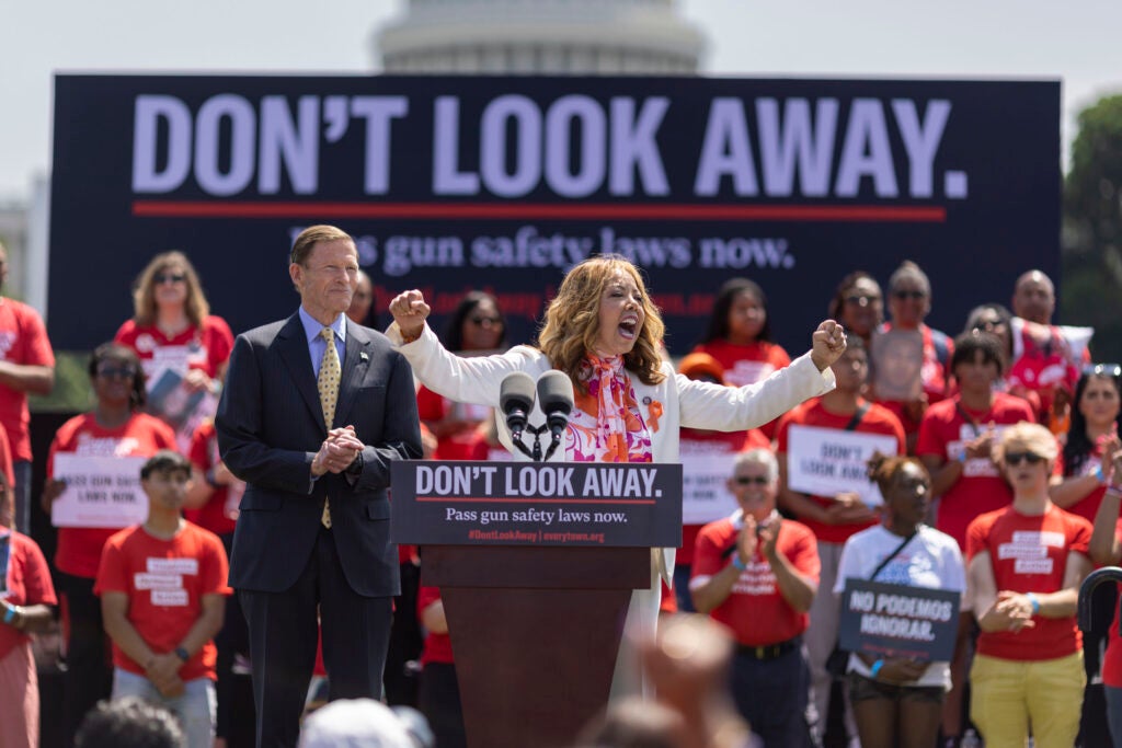 Gun-control advocates gather at a podium beneath a large “Don't Look Away” banner calling for gun safety laws.