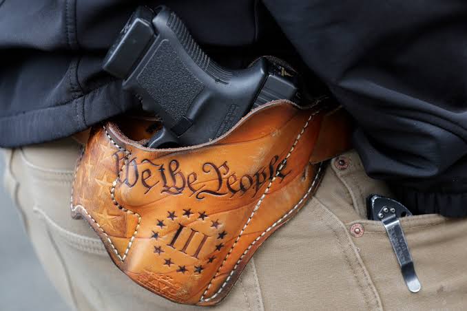 Close-up of a black handgun in a brown leather holster featuring “We the People” and Roman numeral III lettering.